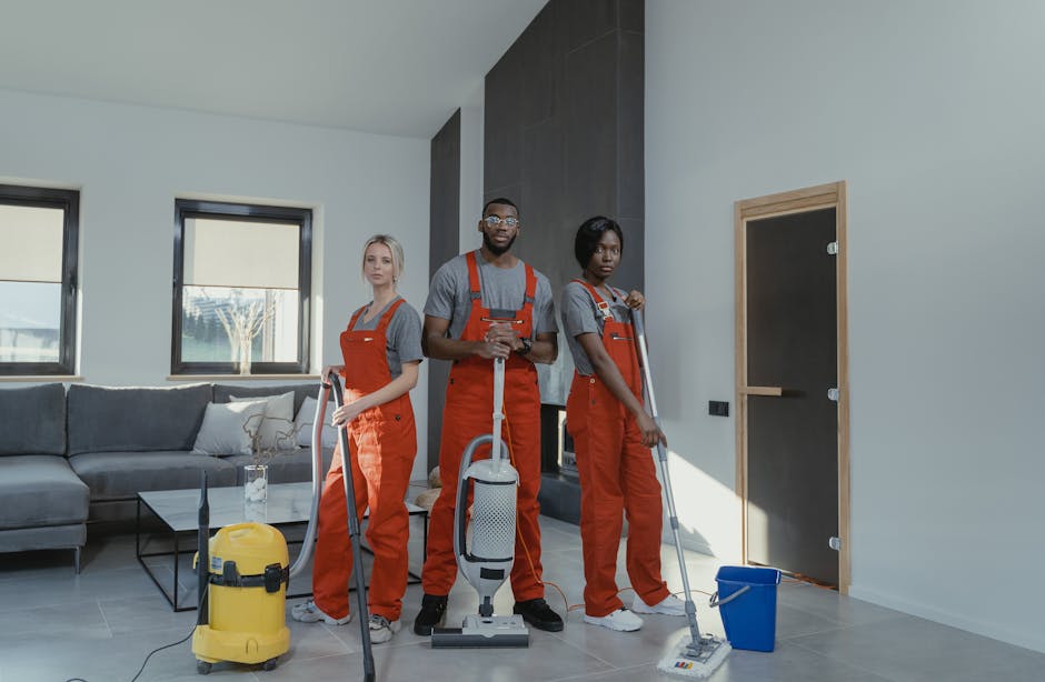 Two professional cleaners from Cleaners Catford engaged in deep surface cleaning of a modern living room. They are using vacuum and mops on the wooden and tiled floors, respectively, to remove dust an