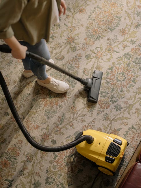 A person cleaning an ornate floral-patterned carpet using a vacuum cleaner with a yellow body and black hose, in a well-lit room. The individual is dressed casually, wearing sneakers, jeans, and a bei